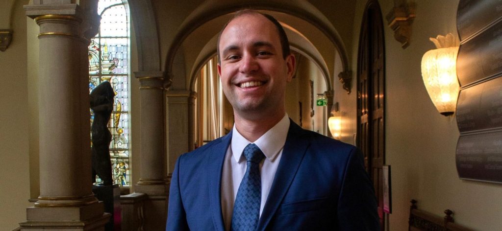 Lars Berndt in een donkerblauw pak in een klassieke gang met bogen en rood tapijt, glimlachend naar de camera.