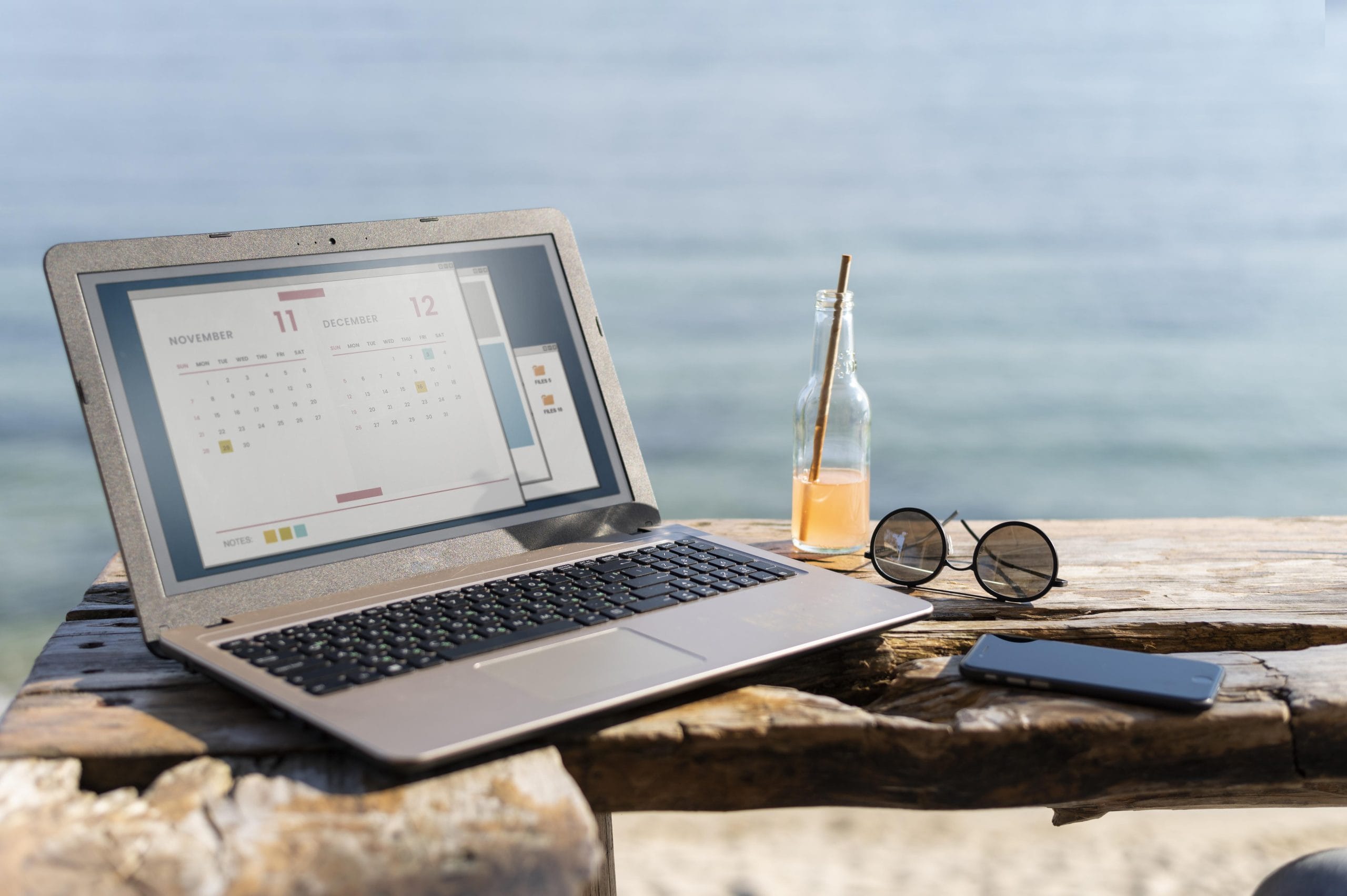 Laptop met een kalender op het scherm, zonnebril en drankje op een houten tafel aan zee.